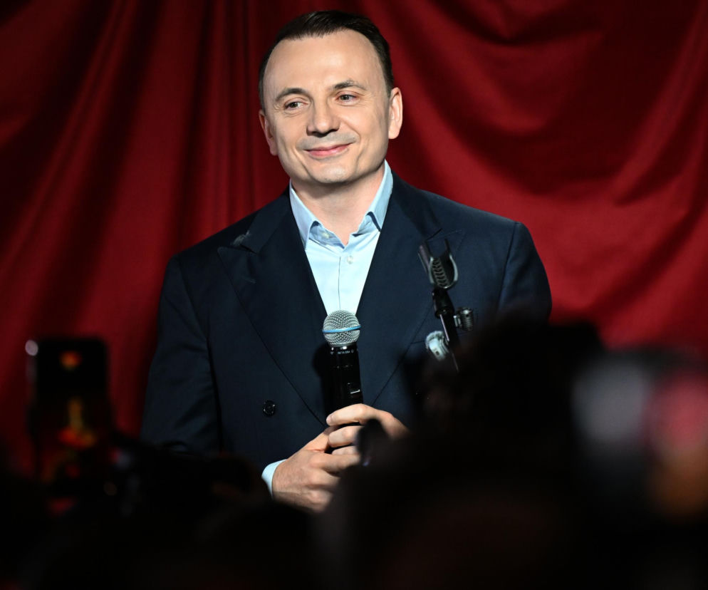 Uśmiechnięty Łukasz Gibała trzyma mikrofon na tle czerwonej kotary. O planach prezydenta Krakowa przeczytasz na SE Polityka.