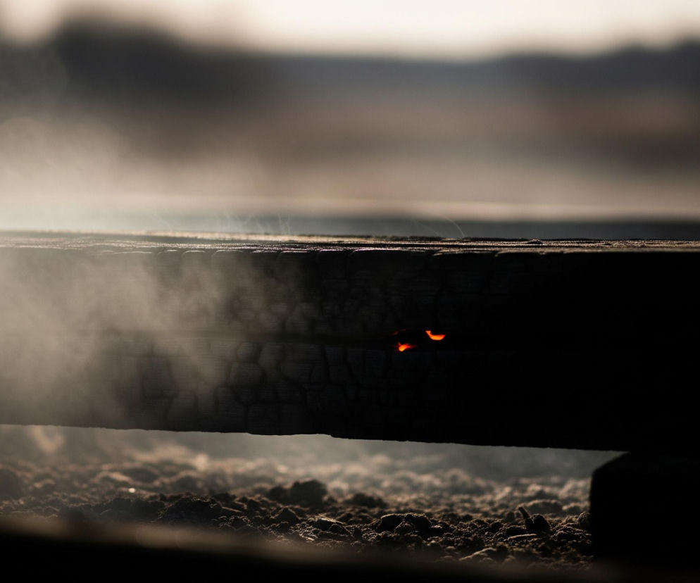 Duży, poczerniały kawałek drewna rozciąga się horyzontalnie przez środek kadru, widoczne są na nim wyraźne pęknięcia i nierówności. Z lewej strony kawałka drewna unosi się biały dym, rozświetlony przez światło padające z tła. Po prawej stronie drewna, w jego ciemnej części, znajdują się dwa małe, jaskrawo pomarańczowe punkty, świadczące o tlących się żarach. Poniżej drewna widać nierówną, ziarnistą powierzchnię w odcieniach brązu i szarości, przypominającą popiół lub ziemię. Górna część zdjęcia jest rozmyta i jasna, tworząc miękkie tło, które stopniowo ciemnieje ku środkowi, podkreślając kontrast z ciemnym drewnem.