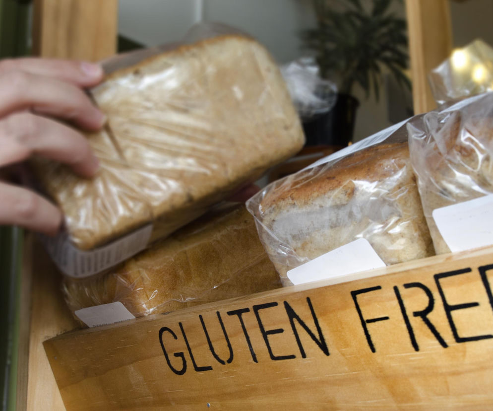 Ręka osoby sięgająca po pakowane pieczywo bezglutenowe w drewnianej skrzynce z napisem GLUTEN FREE. Na Poradnik Zdrowie znajdziesz artykuły ostrzegające przed skutkami ubocznymi mody na dietę bezglutenową bez wskazań medycznych.