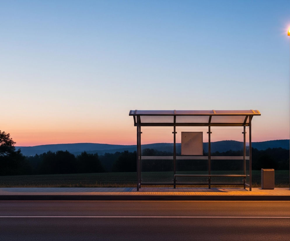 Przystanek autobusowy z przezroczystymi ścianami i dachem, umieszczony po prawej stronie czarnej asfaltowej drogi z białymi liniami. Obok przystanku stoi latarnia uliczna z jasno świecącym, pomarańczowym światłem, oświetlającym część chodnika i wnętrze wiaty. Za przystankiem rozciąga się zielone pole, a dalej ciemne sylwetki drzew i wzgórz, zlewające się z pomarańczowo-niebieskim niebem w tle.