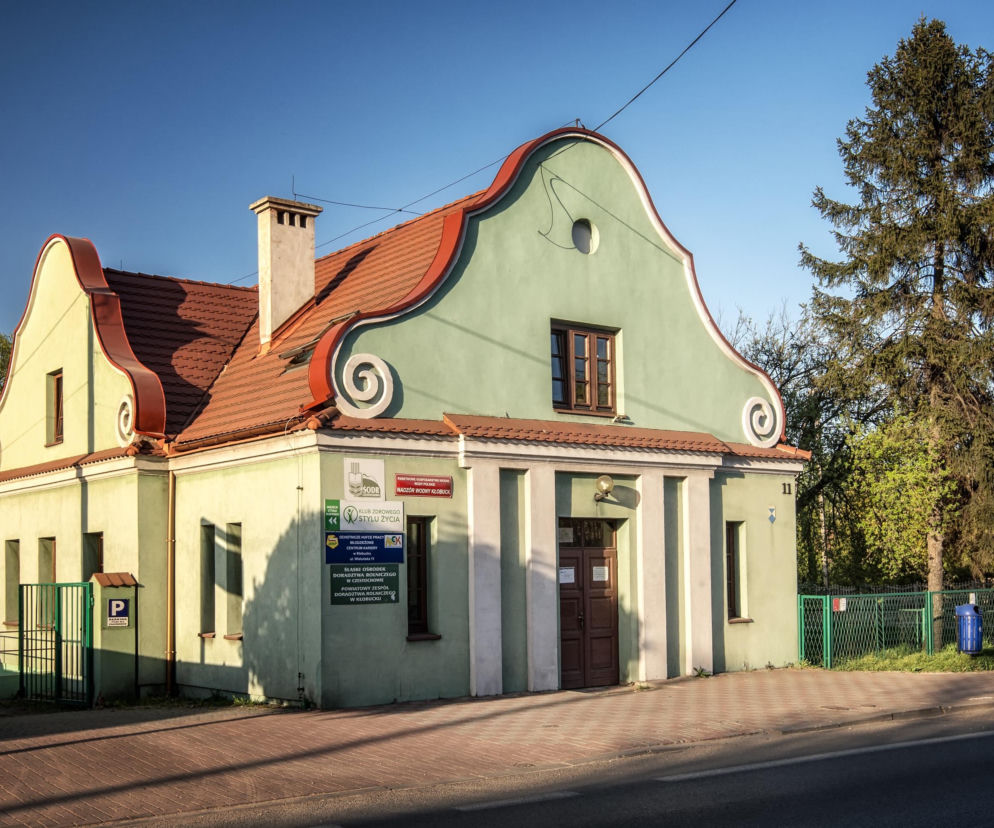 Budynek dawnej łaźni miejskiej w Kłobucku, o jasnozielonej fasadzie i charakterystycznym, falistym dachu z czerwoną dachówką, podkreśla zabytkowy charakter miasta, o którym więcej przeczytasz na naszym portalu. Obiekt stoi przy brukowanym chodniku, widoczne są też tablice informacyjne.