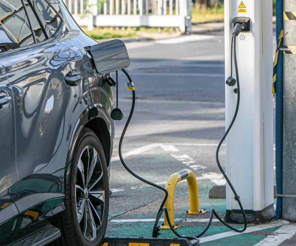 Bok ciemnego samochodu podłączonego kablem do stacji ładowania aut elektrycznych. O badaniu PETRONAS przeczytasz w serwisie Eska.