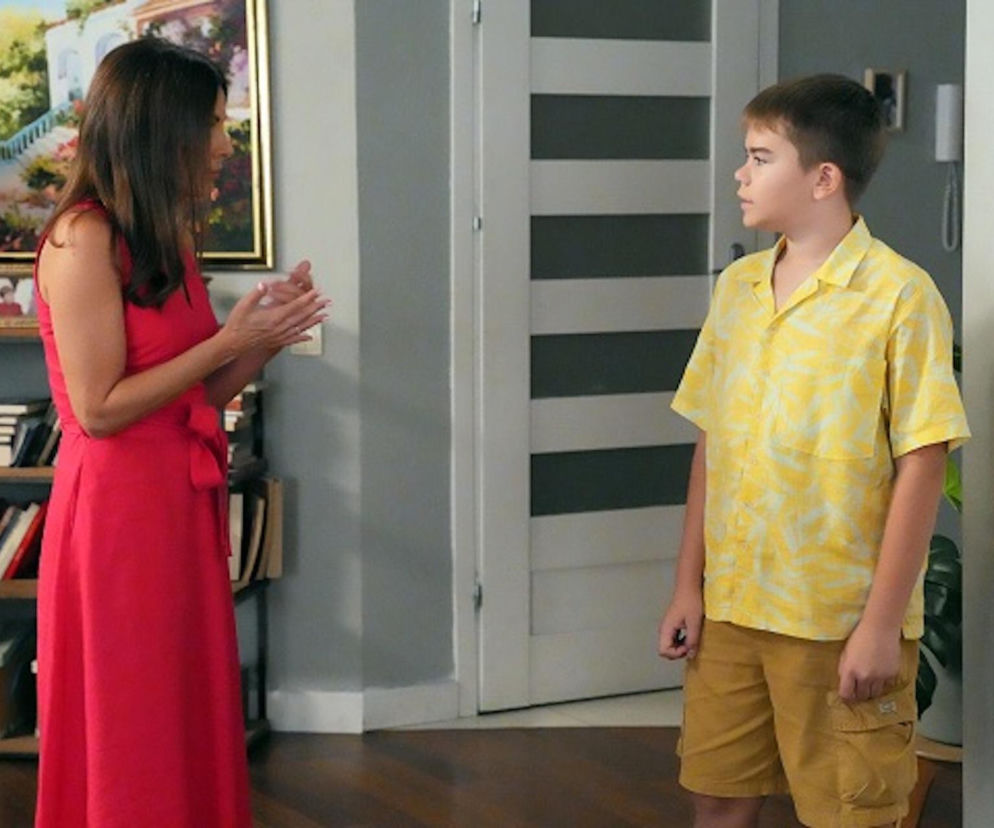 Katarzyna Glinka (Kasia) w czerwonej sukience rozmawia z Bartoszem Gruchotem (Ksawerym) w żółtej koszuli. Matka z synem stoją w pokoju, na tle regału z książkami i obrazu. Opis nagłego zwrotu w życiu Ksawerego oraz desperacji Kasi zwierzącej się Borysowi znajdziesz na Super Seriale.