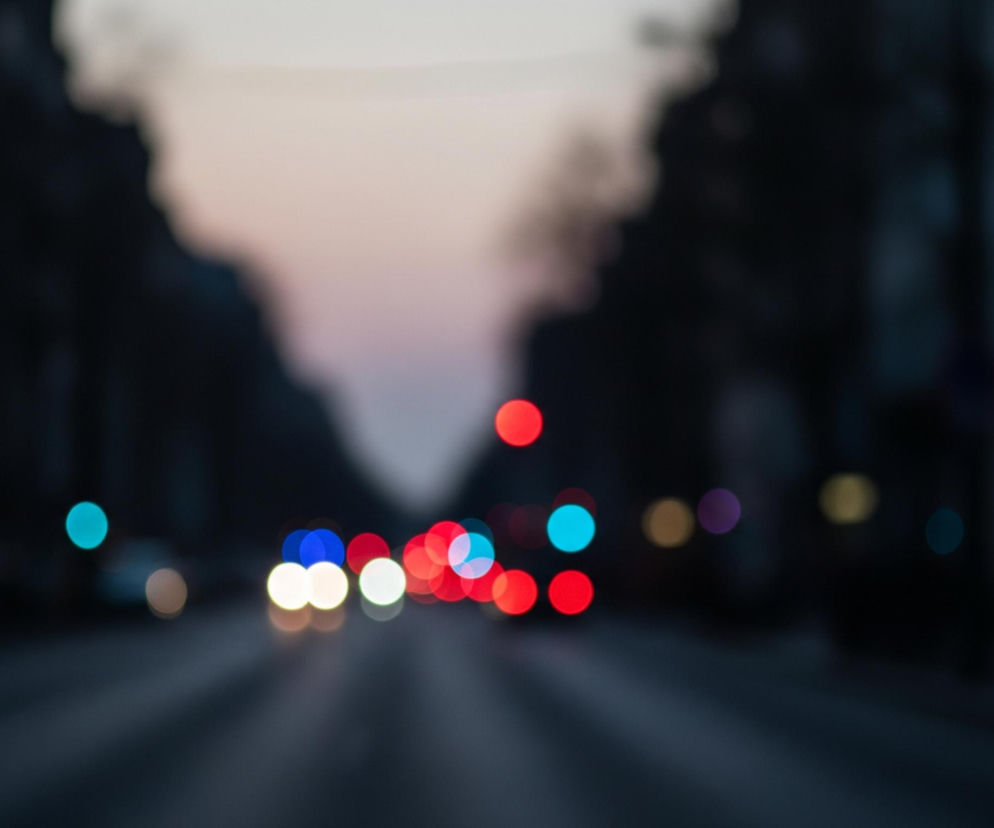 Rozmyty widok ulicy miejskiej o zmierzchu lub w nocy, z licznymi kolorowymi światłami w postaci bokeh. Pośrodku znajduje się grupa jasnych, okrągłych świateł: dominują czerwone, białe oraz dwa niebieskie, sugerujące ruch pojazdów. Droga jest ciemna, a po obu stronach widać niewyraźne, ciemne sylwetki budynków lub drzew. Górna część obrazu ukazuje jaśniejsze, szaro-różowe niebo z przejściem w ciemniejsze odcienie ku krawędziom.
