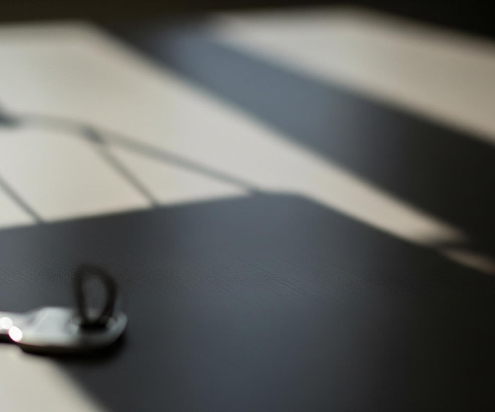 Metalowy klucz leży na ciemnoszarym, teksturowanym blacie, umieszczony w lewym dolnym rogu kadru i częściowo oświetlony jasnym światłem wpadającym z lewej strony. Pośrodku i w prawym górnym rogu blatu dominuje duży, ostro zarysowany cień o skomplikowanym geometrycznym kształcie, sugerujący obecność jakiegoś obiektu poza kadrem. Cień ten przechodzi od jasnego szarości w górnej części do ciemnego szarości i czerni w prawej części zdjęcia, tworząc kontrastujące pasy światła i cienia.