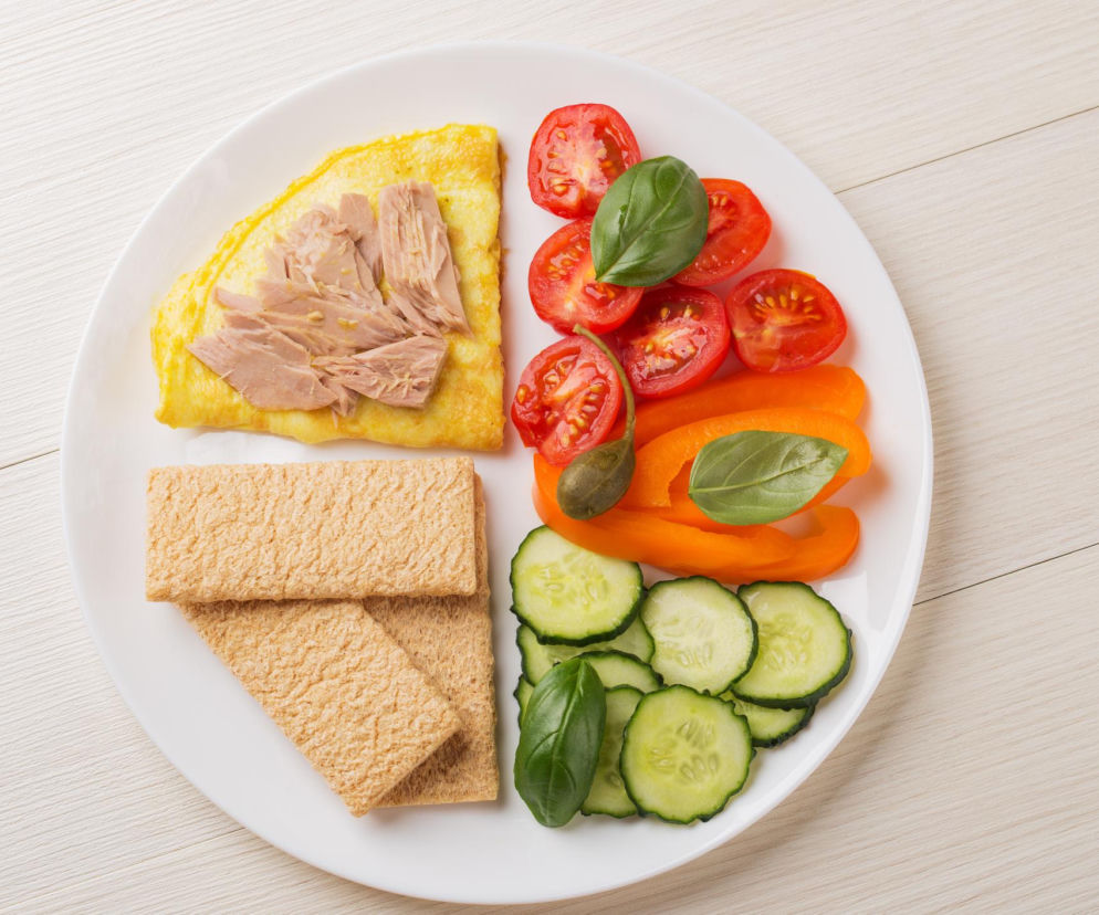 Talerz z jedzeniem, widok z góry. Na białym talerzu znajduje się omlet z tuńczykiem, pomidorki koktajlowe, pomarańczowa papryka, ogórek, liście bazylii oraz pieczywo chrupkie. Poszczególne składniki ułożono w celu wskazania odpowiedniej kolejności jedzenia, by trzymać cukier w ryzach i wesprzeć trzustkę. Więcej o diecie dla osób z insulinoopornością przeczytasz na Poradnik Zdrowie.