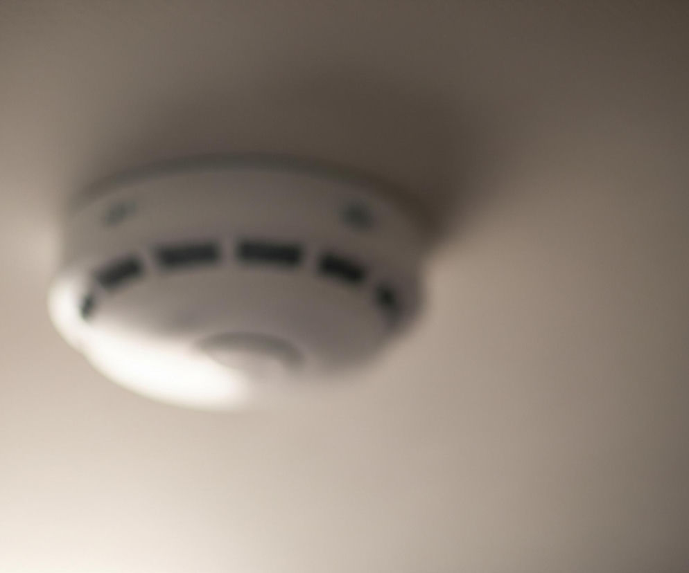 Na suficie, dominującym odcieniem beżu, zamontowany jest okrągły, biały czujnik dymu. Ma on cylindryczny kształt z lekko zaokrągloną kopułą, z kilkoma równomiernie rozmieszczonymi prostokątnymi otworami wokół obwodu. Cały obraz jest nieostry, z centralnym punktem czujnika nieco wyraźniejszym, a tłem bardziej rozmytym, szczególnie w prawym górnym rogu. Światło pada z lewej strony, tworząc delikatny cień pod czujnikiem.