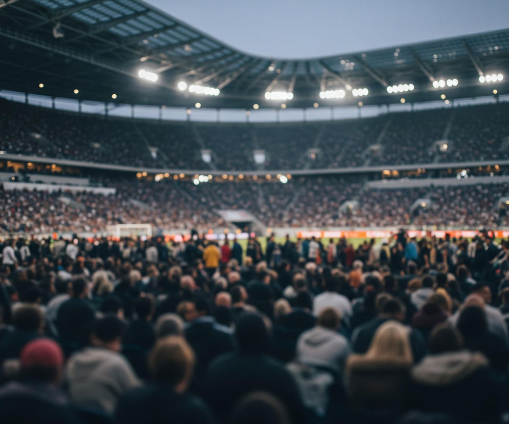 Rozległy stadion sportowy wypełniony tłumem widzów rozciąga się od pierwszego planu po horyzont. Ostrość obrazu skupia się na plecach osób siedzących na pierwszym planie, ubranych w ciemne kurtki i bluzy, z pojedynczymi akcentami kolorystycznymi, takimi jak czerwona czapka. Dalej, w gęsto zaludnionych rzędach trybun, widać znacznie mniejsze, rozmyte sylwetki ludzi, tworzące mozaikę białych, szarych i czarnych punktów. Górna część stadionu, wraz z dachem i jaskrawo świecącymi reflektorami, rozświetla wieczorne, błękitno-szare niebo.