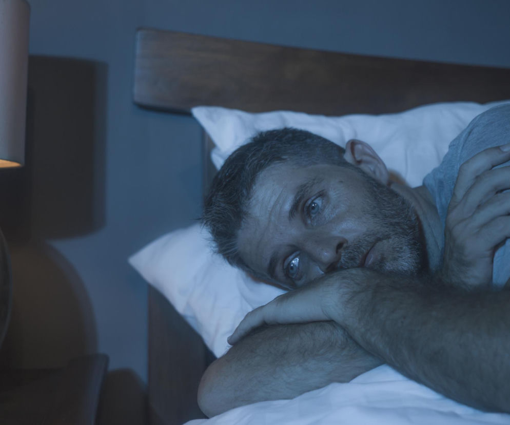 Mężczyzna z bezsennością leży w łóżku, spoglądając w stronę lampki nocnej, co symbolizuje problemy z zasypianiem. Jego zmartwiona twarz i błękitne światło tworzą atmosferę niepokoju. Na Poradnik Zdrowie znajdziesz wskazówki, jak poprawić jakość snu.