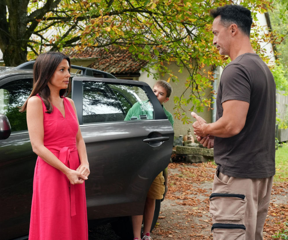 Katarzyna Glinka w różowej sukience stoi obok samochodu, rozmawiając z Rafałem Cieszyńskim w brązowej koszulce i beżowych spodniach. W tle widać dziecko, Ksawerego, wyglądające zza otwartych drzwi auta. Scena konfliktu emocjonalnego bohaterów serialu Barwy szczęścia na Super Seriale.