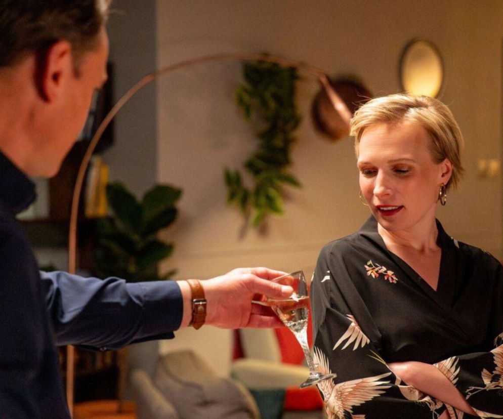 Sonia (Weronika Nockowska) w czarnej, kwiecistej bluzce z szerokimi rękawami, opuszcza wzrok, gdy Stefaniak (Paweł Ławrynowicz) w ciemnej koszuli podaje jej kieliszek szampana. Scena z serialu Barwy szczęścia dostępnego na platformie Super Seriale.