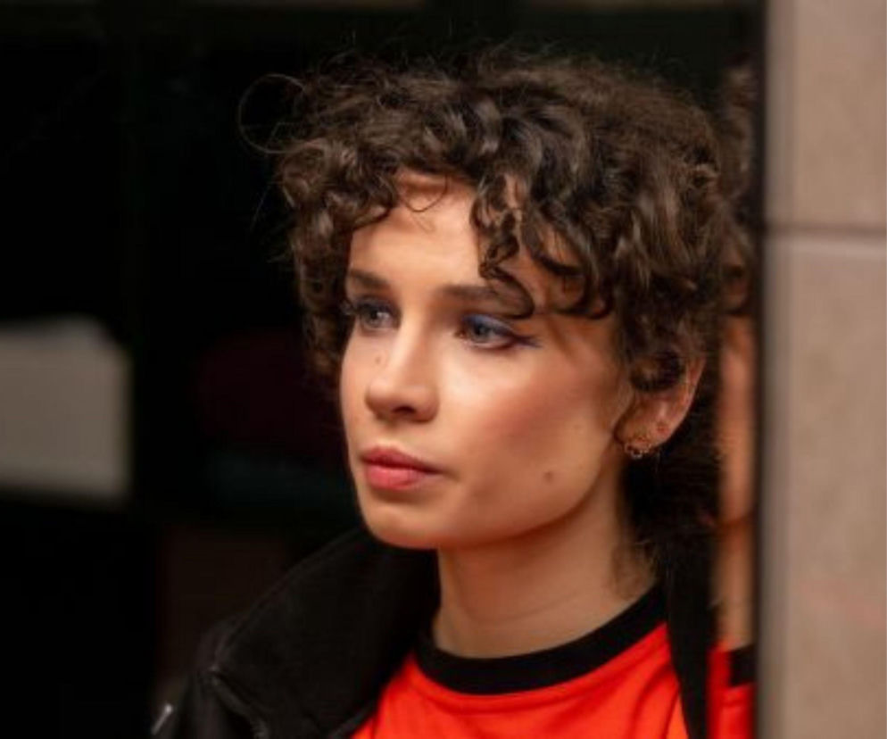 Kobieta o kręconych, ciemnych włosach i intensywnym, niebieskim makijażu oczu, ubrana w czarną kurtkę na pomarańczową koszulkę, wpatrująca się zamyślona w dal. Ta postać może być jedną z bohaterek najnowszego odcinka serialu Na sygnale, o którym więcej przeczytasz na portalu Super Seriale.