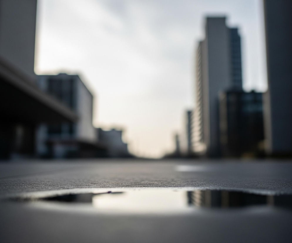 Na pierwszym planie, w dolnej części obrazu, znajduje się mała kałuża wody o nieregularnym kształcie, odbijająca jasne niebo i zamazane fragmenty budynków w oddali. Powierzchnia wokół kałuży to ciemnoszary, teksturowany chodnik lub asfalt, który stopniowo jaśnieje ku górze i jest rozmyty. W tle widoczne są niewyraźne, wysokie budynki biurowe w odcieniach szarości i granatu, otoczone jasnym, nieco zachmurzonym niebem.