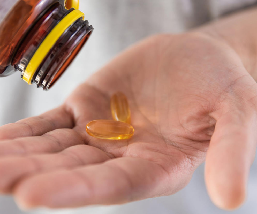 Dłoń osoby dorosłej trzymająca dwie bursztynowe kapsułki z suplementem diety, prawdopodobnie witaminą D, wysypane z brązowej butelki. Zdjęcie ilustruje temat prawidłowego przyjmowania witaminy D i jej wpływu na zdrowie, o czym przeczytasz na portalu Poradnik Zdrowie.