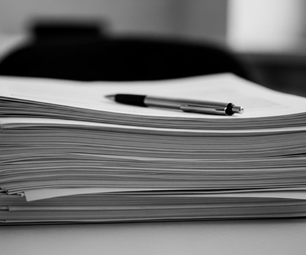 Na pierwszym planie, centralnie, leży stos papierów w odcieniach szarości, ułożonych równo jeden na drugim. Na wierzchu stosu leży długopis z metalową, błyszczącą obudową i czarnym, gumowym uchwytem, skierowany w prawo. Papiery spoczywają na jasnoszarym, lekko teksturowanym blacie. W tle, rozmyte i ciemne, widać zarys mebli lub wyposażenia biurowego, w tym fragmenty ciemnego, niemal czarnego krzesła i jaśniejszych ścian.