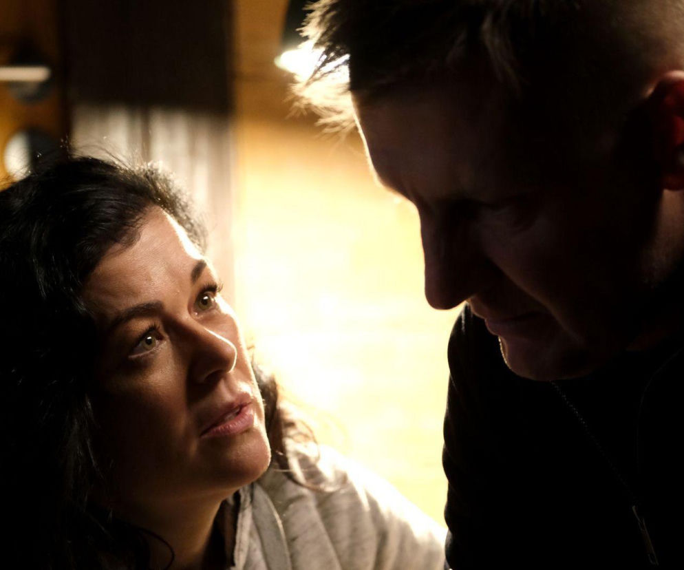 Kinga (Katarzyna Cichopek) i Piotrek (Marcin Mroczek) w zbliżeniu, rozmawiający ze sobą w słabym oświetleniu, co oddaje dramatyczną atmosferę odcinka M jak miłość. Tę i inne informacje o serialach znajdziesz na Super Seriale.