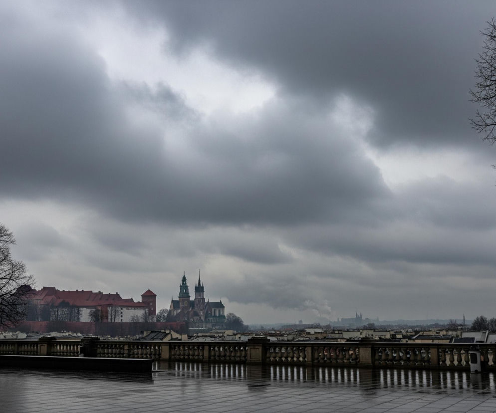 Na pierwszym planie widoczny jest mokry, ciemny chodnik z ułożonych kwadratowych płyt, odbijający światło. Po jego długości ciągnie się kamienna balustrada z kolumienkami, również odbijająca jasne refleksy. Dalej, za balustradą, rozciąga się panorama miasta. Na horyzoncie dominują historyczne budowle z czerwonymi dachami, a centralnie wyróżniają się wieże kościoła z ostrymi spiczastymi zakończeniami. Po lewej stronie kadru stoi duże, bezlistne drzewo, którego gałęzie odbijają się w mokrej nawierzchni, a po prawej stronie znajduje się fragment podobnego drzewa. Niebo jest pochmurne i szare, z jaśniejszymi fragmentami chmur.