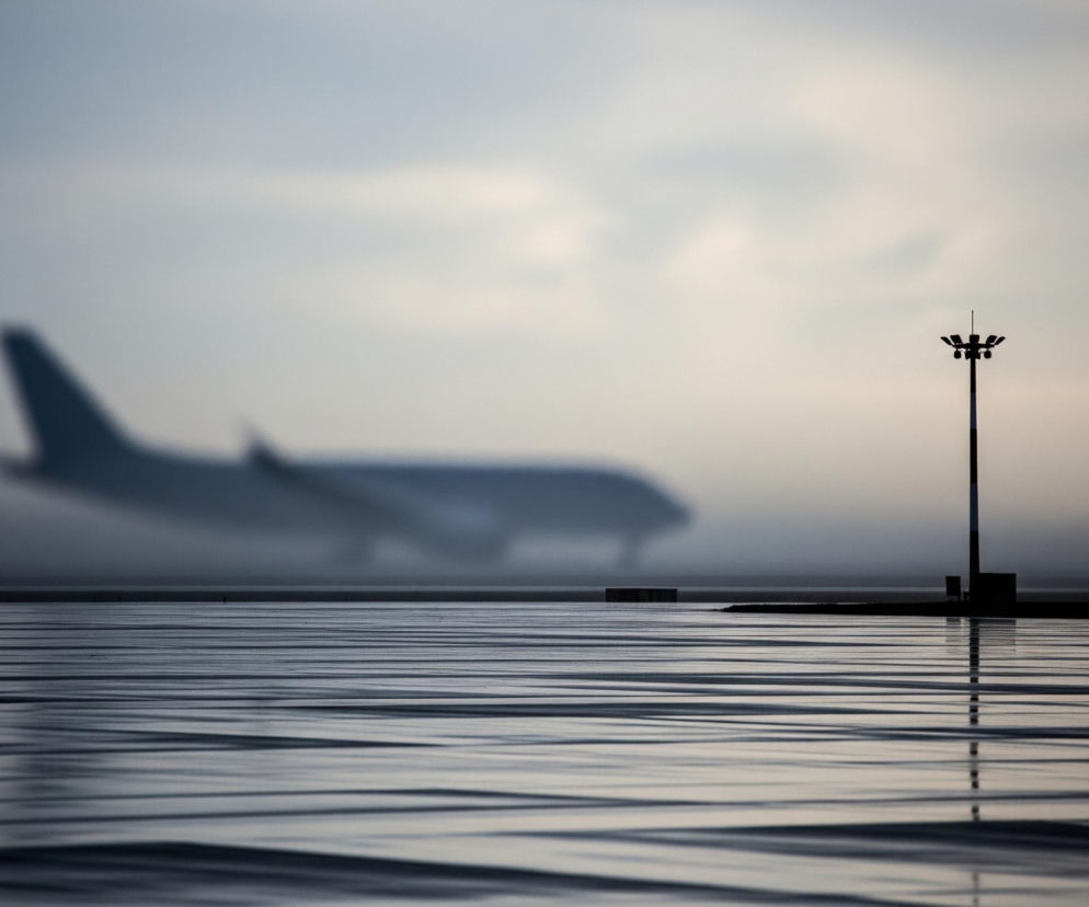 Na pierwszym planie, z prawej strony, stoi wysoki czarny słup oświetleniowy z kilkoma reflektorami skierowanymi w różne strony, widocznymi w górnej części. Powierzchnia pod nim, zajmująca dolną połowę obrazu, jest lśniąca i poziomo pofalowana, odbijając światło w postaci równoległych, jasnych i ciemnych pasów. W tle, niewyraźny kontur dużego samolotu pasażerskiego, ciemniejszego niż niebo, jest umieszczony po lewej stronie, poruszając się w prawo. Całe tło stanowi zamglone, jasne niebo w odcieniach szarości i błękitu, co nadaje scenie enigmatyczny i spokojny nastrój.