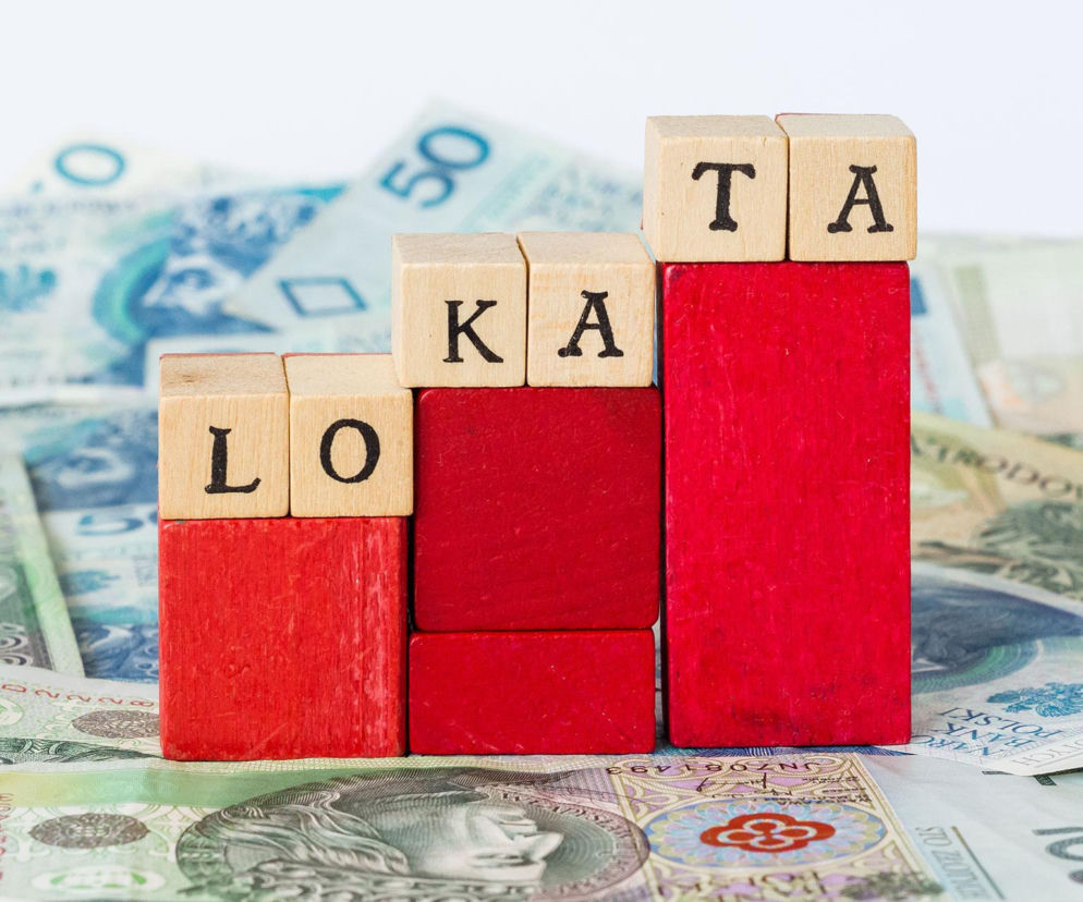 Drewniane klocki z literami układające się w słowo „LOKATA”, umieszczone na czerwonych podwyższeniach przypominających wykres słupkowy. W tle rozłożone są banknoty o nominałach 50 i 100 złotych, symbolizujące oszczędności i zyski. Dowiedz się więcej o lokatach na Super Biznes.