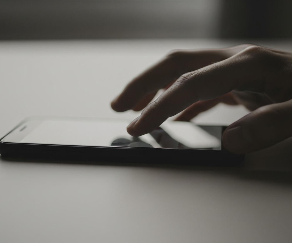 Czarny smartfon z jasnym ekranem leży poziomo na gładkiej, jasnoszarej powierzchni. Ciemna ręka z częściowo zgiętymi palcami dotyka ekranu, a dwa palce są widoczne w górnej części. Tło jest ciemnoszare i rozmyte, z niewyraźnym przejściem kolorów od jasnoszarego do ciemnoszarego w górnej części kadru.