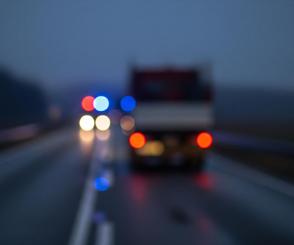 Rozmazany widok przedstawia drogę o zmierzchu lub w nocy, z niewyraźnie widoczną ciężarówką na pierwszym planie, jej tył jest skierowany do widza. Światła ciężarówki to dwie czerwone, okrągłe plamy. Nad ciężarówką i po jej lewej stronie widoczne są rozmyte punkty świetlne o kolorach czerwonym, błękitnym i białym. Nawierzchnia drogi jest ciemna, a jej prawy pas ma białe, rozmazane linie. Tło składa się z ciemnych, niewyraźnych kształtów przypominających drzewa i nieba w odcieniach szarości i błękitu.