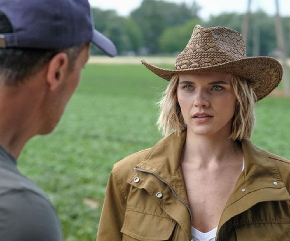 Janka (Oliwia Drożdżyk) w kapeluszu i brązowej kurtce patrzy z niepokojem na Mariusza (Rafał Cieszyński) odwróconego plecami, w szarej koszulce i granatowej czapce, na tle zielonego pola. Scena z odcinka „Barw szczęścia” o kłótni o kapelusz Marleny. Więcej na Super Seriale.