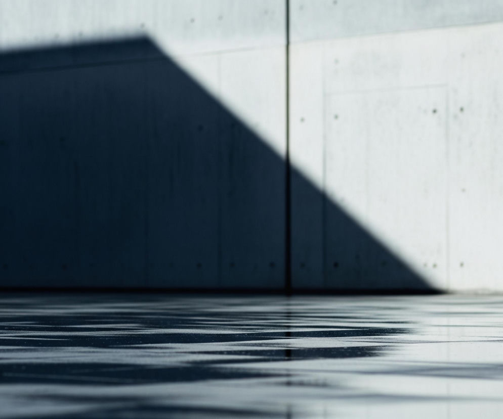 Skośny cień o ostrych krawędziach dzieli pionową, jasnoszarą ścianę na dwie części: lewą, pogrążoną w ciemności, i prawą, oświetloną. Dolna krawędź cienia rozciąga się również po poziomej powierzchni podłogi, tworząc kontrastujące pasy światła i mroku. Lewa strona ściany, znajdująca się w cieniu, jest ciemnoniebieska, natomiast prawa jest jasnoszara, z widocznymi drobnymi plamkami i ryskami na powierzchni. Na podłodze widoczne są nieregularne, faliste refleksy światła, przypominające odbicia na wodzie lub chropowatą teksturę, które mieszają się z obszarami w cieniu. Pionowa linia dzieli ścianę na dwie równe sekcje.