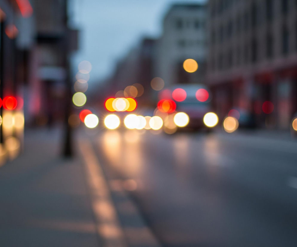 Ulica w mieście o zmierzchu lub w nocy, sfotografowana z efektem bokeh, co sprawia, że światła są rozmyte i tworzą kolorowe kręgi. Po lewej stronie widać chodnik, a dalej budynki, z których odbijają się ciepłe, żółte światła. Ulica po prawej stronie wypełniona jest rozmytymi światłami samochodów – dominują żółte, białe i czerwone plamy, wskazujące na ruch pojazdów. Tło stanowią niewyraźne zarysy budynków pod lekko zachmurzonym, szarawym niebem.