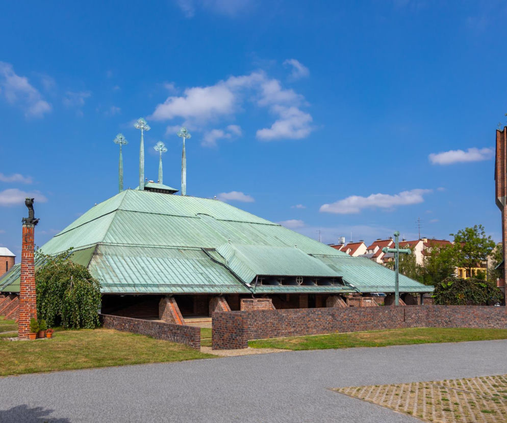 Kościół Świętego Ducha w Tychach z zielonym dachem o geometrycznym kształcie, ceglaną fasadą i smukłą dzwonnicą, zaprojektowany przez Stanisława Niemczyka, na tle błękitnego nieba z białymi chmurami. O architekturze sakralnej, w tym o tym kościele, przeczytasz na Architektura Murator Plus.