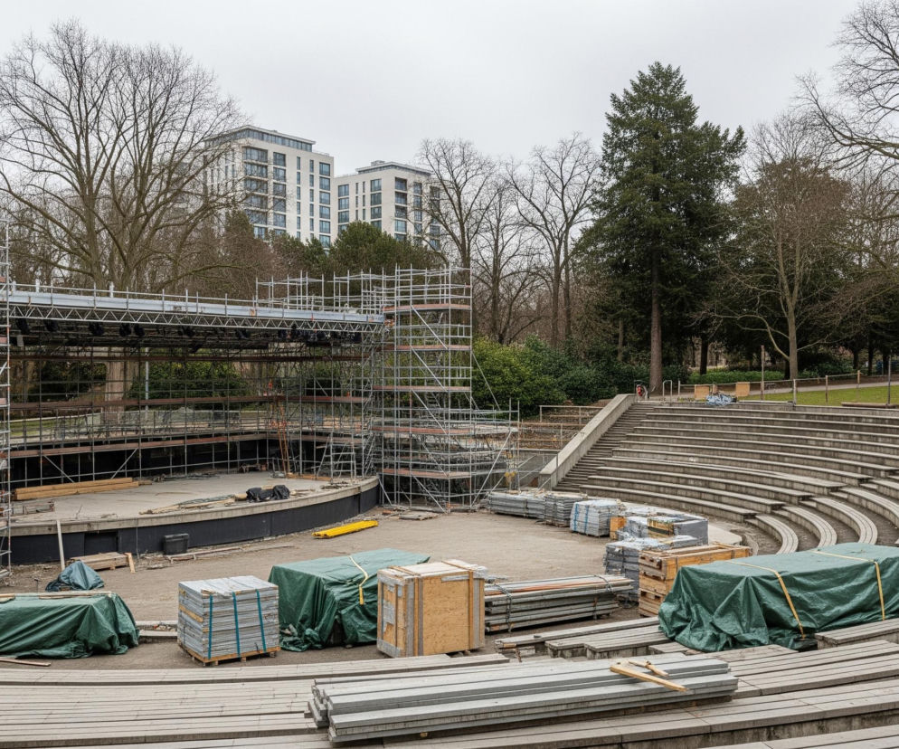 Otwarty amfiteatr w trakcie budowy lub demontażu, ze sceną z lewej strony pokrytą rusztowaniem, a widownią z betonowymi ławkami po prawej. Na scenie leżą materiały budowlane, a wzdłuż pierwszych rzędów widowni piętrzą się stosy desek, skrzynie oraz opakowania przykryte zieloną plandeką, gotowe do użycia lub transportu. W tle za amfiteatrem znajdują się bezlistne drzewa i kilka nowoczesnych budynków mieszkalnych o jasnych elewacjach, a na horyzoncie widoczne jest zachmurzone, szare niebo.