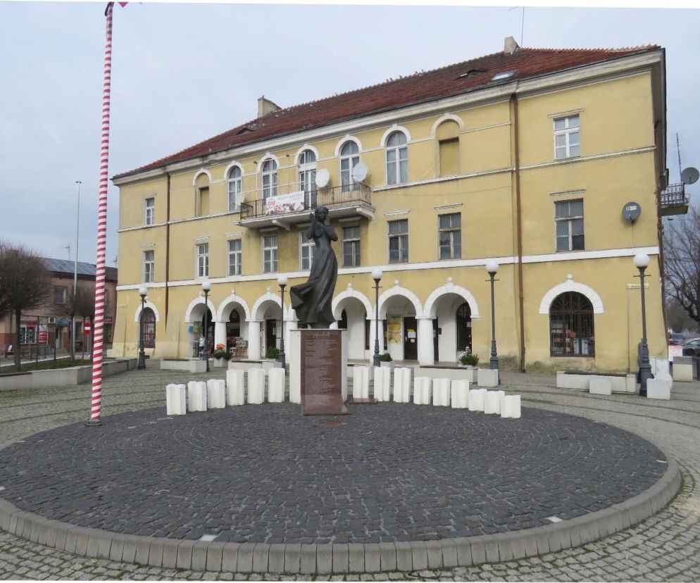 Rynek w Błaszkach z pomnikiem i zabytkowym, jasnym budynkiem z arkadami oraz balkonem, ukazującym historyczną architekturę miasta. Więcej o tym urokliwym miasteczku w woj. łódzkim przeczytasz na naszym portalu.