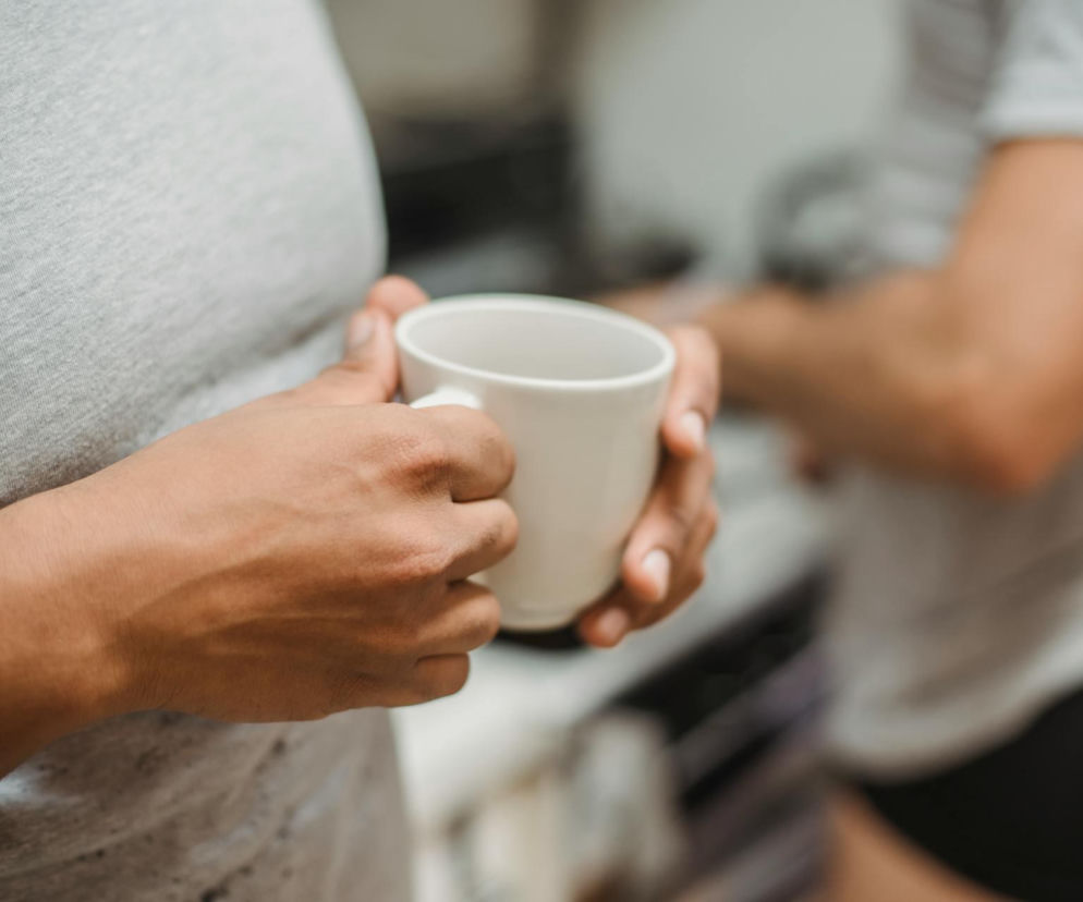 Osoba trzymająca biały kubek kawy, w tle niewyraźna postać. Artykuł na Poradnik Zdrowie wyjaśnia, jak bezpiecznie odstawić kawę i uniknąć bólu głowy, omawiając skutki nagłego detoksu kofeinowego i sposoby zmniejszania objawów odstawiennych.