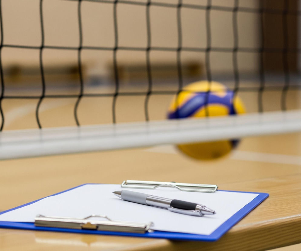 Na pierwszym planie, z prawej strony, leży niebieska podkładka z przypiętym do niej białym arkuszem papieru, a na papierze spoczywa srebrno-czarny długopis. Obiekt ten leży na jasnobrązowej, drewnianej powierzchni. Za tym znajduje się czarna siatka o dużych oczkach, rozciągająca się przez całą szerokość kadru, z białym, poziomym pasem u dołu. Po lewej stronie, za siatką, niewyraźnie widoczna jest żółto-niebieska piłka do siatkówki, a tło stanowią rozmyte, jasne powierzchnie sali sportowej.