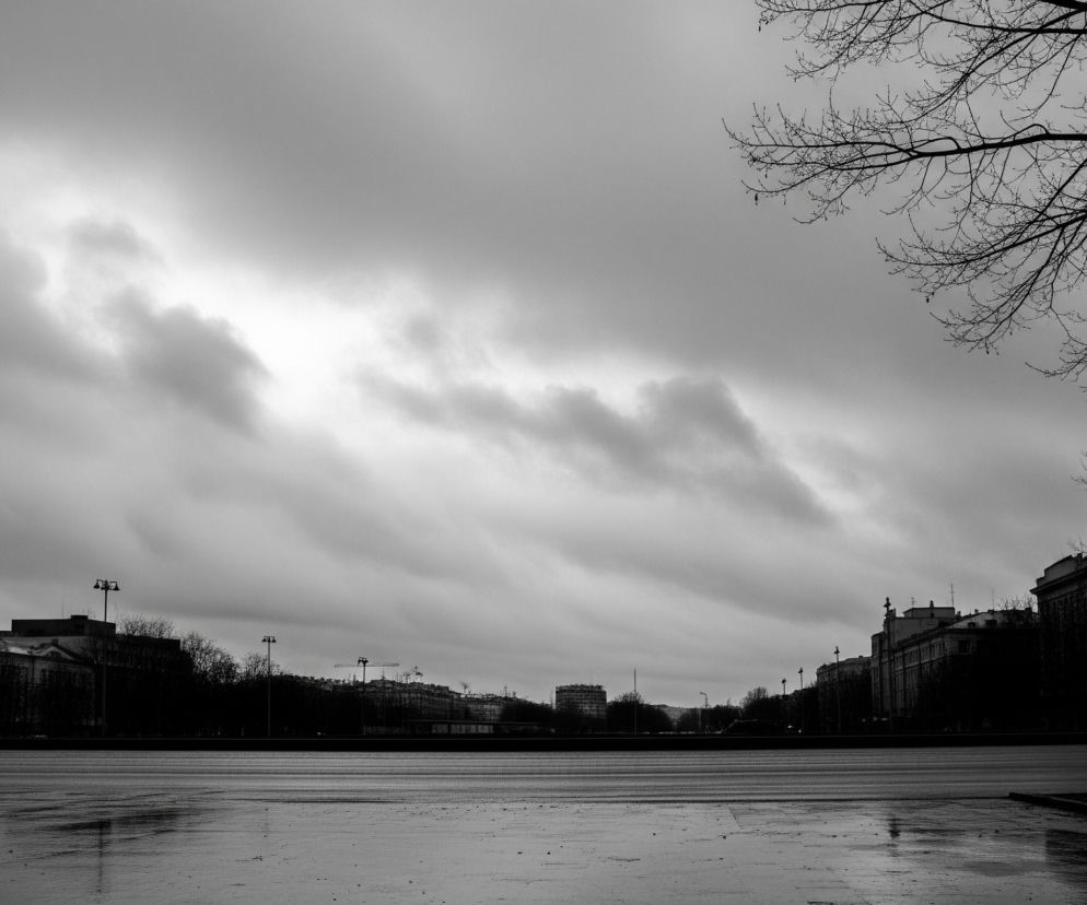 Czarno-białe zdjęcie przedstawia miejski krajobraz z nisko zawieszonym, zachmurzonym niebem, które zajmuje większą część kadru. Na pierwszym planie widoczna jest mokra powierzchnia, przypominająca chodnik lub plac, z licznymi odbiciami światła. Wzdłuż horyzontu rozciągają się budynki i drzewa, tworzące ciemne sylwetki. Po prawej stronie kadru, w górnej części, widoczne są nagie gałęzie drzewa.