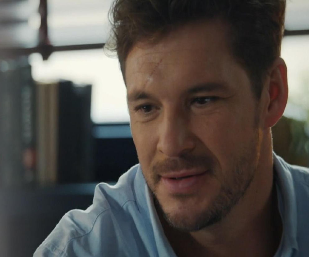 Mikołaj Roznerski jako Marcin Chodakowski w scenie z serialu M jak miłość. Mężczyzna ma krótkie, ciemne włosy, widoczną bliznę na czole i delikatny zarost. Ubrany jest w jasnoniebieską koszulę. Wygląda na zamyślonego, jego wzrok skierowany jest w dół. Więcej informacji o serialach, takich jak M jak miłość, znajdziesz na Super Seriale.