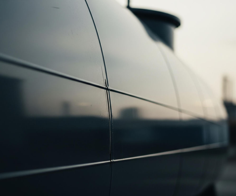 Na pierwszym planie, z lewej strony, widoczna jest duża, metaliczna powierzchnia w ciemnych odcieniach szarości, niemal czerni, z poziomymi i pionowymi liniami podziału. Powierzchnia ta, prawdopodobnie fragment dużego zbiornika lub pojazdu, jest lekko zakrzywiona, a na jej odbijającej światło płaszczyźnie widać rozmyte refleksy otoczenia. W górnej części kadru, po prawej stronie, majaczy ciemny, niezidentyfikowany obiekt wystający ponad główną konstrukcję. Tło jest rozmyte i jasne, w odcieniach szarości i bieli, z niewyraźnymi konturami budynków i jasnym punktem, być może słońcem, lub lampą, w prawym środku.