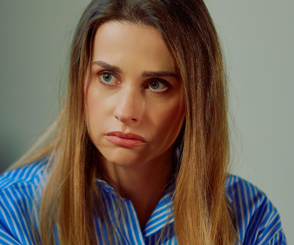 Eliza Bergowa z serialu Na Wspólnej w zbliżeniu, ma długie, rozpuszczone włosy, a jej twarz wyraża zmartwienie. Kobieta ubrana jest w niebieską koszulę w białe paski. Ten dramatyczny wątek można śledzić na portalu Super Seriale.