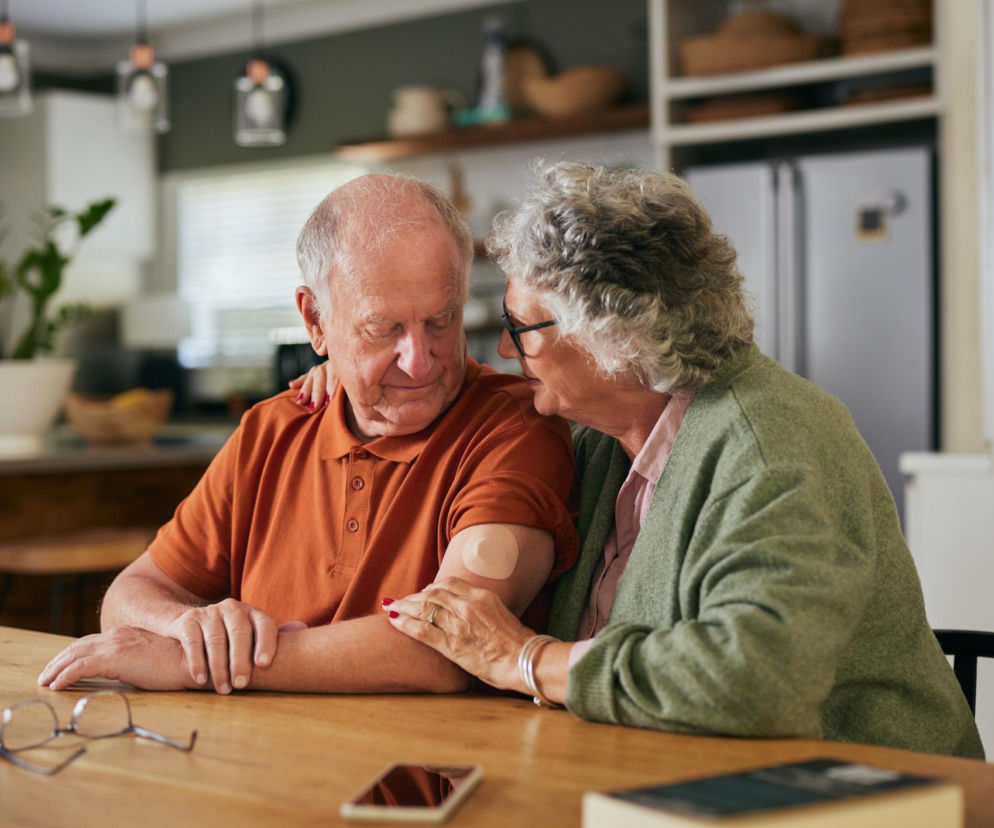 Starsza kobieta obejmuje ramieniem starszego mężczyznę siedzącego przy stole, na którego ramieniu widać plaster. Obydwoje spoglądają na siebie z troską, co symbolizuje wsparcie w leczeniu cukrzycy typu 2. Więcej o statynach i diabetykach przeczytasz na Poradnik Zdrowie.