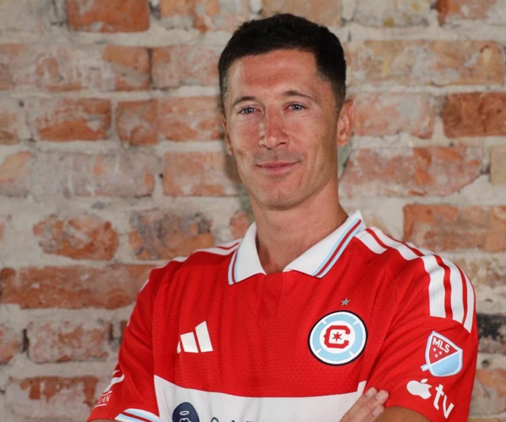 Robert Lewandowski w koszulce Chicago Fire z logo MLS na ramieniu i herbem klubu na piersi, uśmiecha się, mając skrzyżowane ręce na tle ceglanej ściany. O jego kosmicznych zarobkach przeczytasz więcej na naszym portalu.