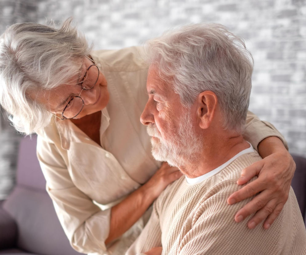 Starsza kobieta w beżowej koszuli obejmująca mężczyznę z siwą brodą, symbolizująca wsparcie i opiekę. Obraz odzwierciedla troskę o bliskich, nawiązując do wyzwań związanych z chorobą Alzheimera. Więcej o wczesnej diagnostyce przeczytasz na Poradnik Zdrowie.