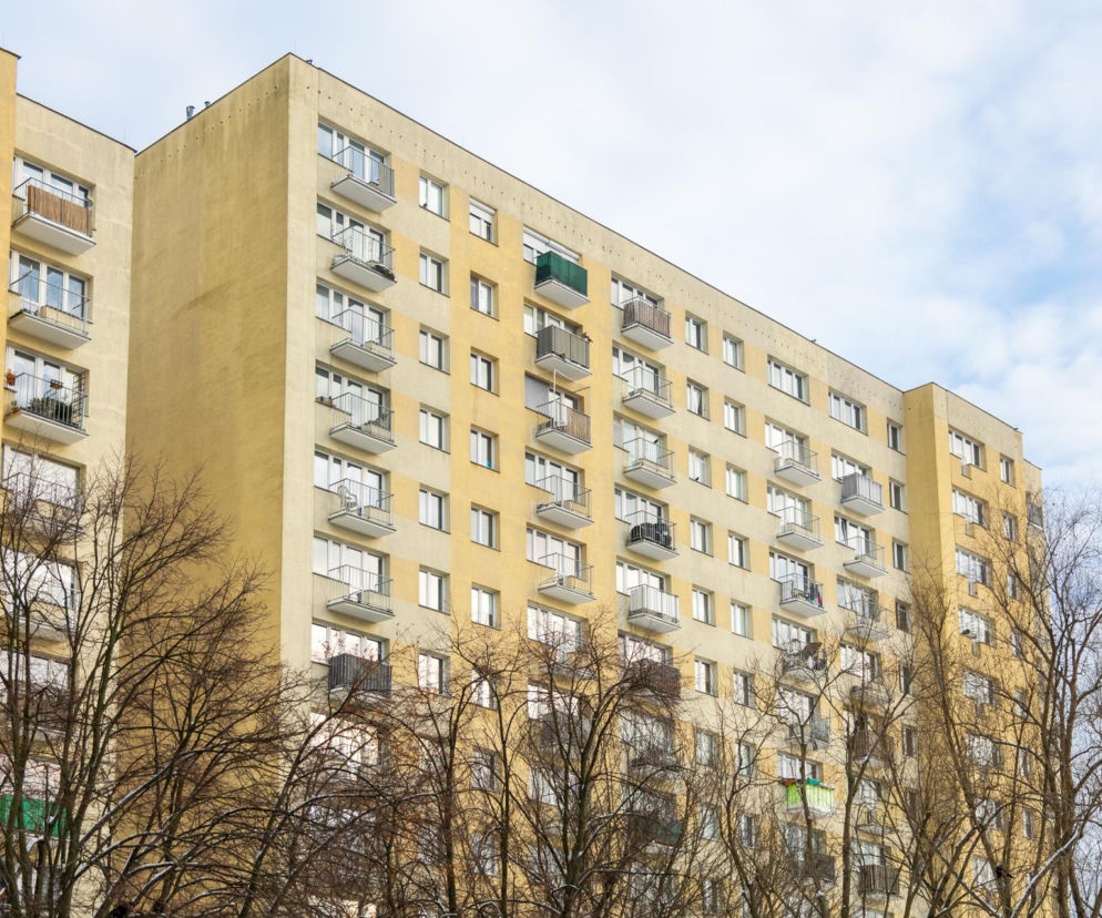 Żółty blok mieszkalny z licznymi balkonami, typowy dla budownictwa wielkiej płyty w Warszawie, widoczny pod pochmurnym niebem. Drzewa bez liści na pierwszym planie symbolizują dyskusję o zmianach w prawie dotyczącym mieszkań komunalnych na Super Biznes.