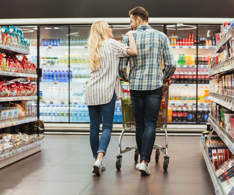 Para młodych ludzi z wózkiem na zakupach w supermarkecie. Kobieta z długimi blond włosami i mężczyzna w koszuli w kratę idą wzdłuż alejek z produktami, przypominając, że informacje o niedzielach handlowych znajdziesz na Super Biznes.