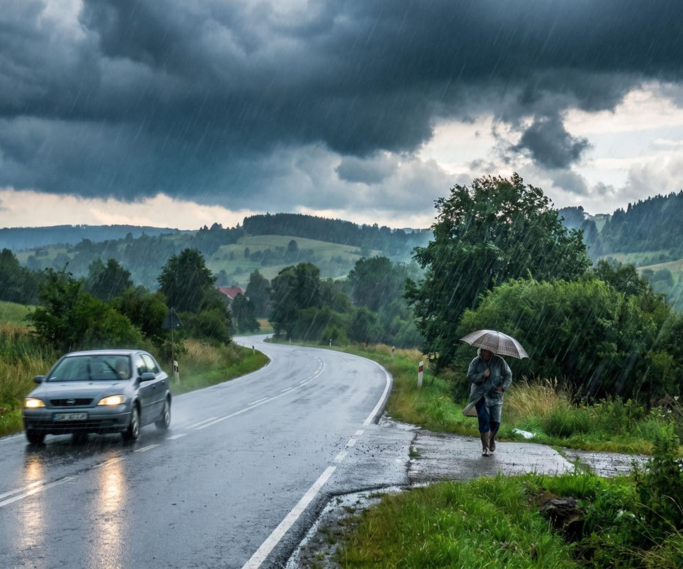 Gwałtowna ulewa zalewająca asfaltową drogę wijącą się przez zielone wzgórza z lasami. Na pierwszym planie idąca osoba z parasolem, a obok niej jadące auto z włączonymi światłami. Burze na południu Polski mogą przynieść ulewne opady i silny wiatr, o czym przeczytasz na naszym portalu.
