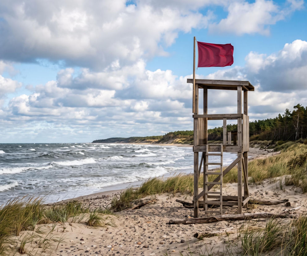 Drewniana wieża ratownicza z czerwoną flagą na pustej plaży. O zakazach kąpieli w Mielnie przeczytasz na SE Szczecin.
