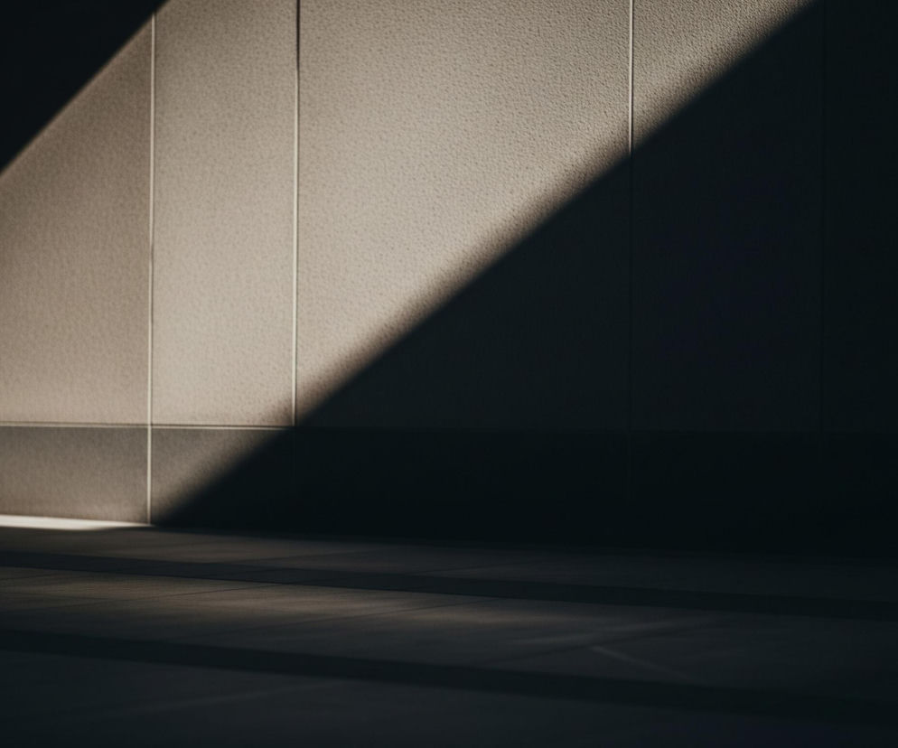 Światło dzienne pada ukośnie z lewego górnego rogu na teksturowaną ścianę, tworząc wyraźny trójkąt jasnej powierzchni w górnej lewej części obrazu. Ściana, podzielona na pionowe i poziome prostokąty cienkimi fugami, ma szorstką, ziarnistą fakturę w odcieniach beżu i jasnego brązu w oświetlonych miejscach. Pozostała część obrazu pogrążona jest w cieniu, ujawniając jedynie ciemne, ledwie widoczne detale podłogi i dolnej części ściany, a także drobne odbicia światła na horyzontalnej krawędzi.