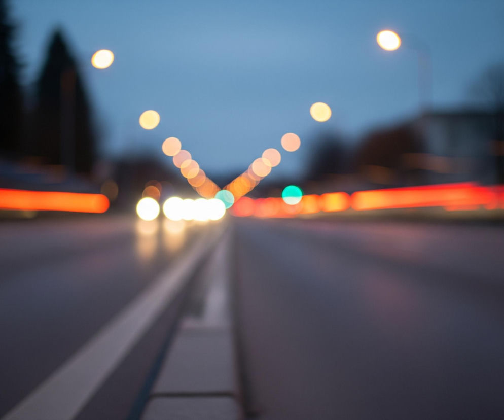 Rozmazany obraz przedstawia nocną ulicę lub autostradę z światłami samochodów i latarni. Centralnym punktem jest ciemna asfaltowa droga z wyraźną białą linią oddzielającą pasy ruchu po lewej stronie i szarym, betonowym krawężnikiem pośrodku. Po obu stronach drogi rozciągają się rozmyte smugi czerwonego światła, sugerujące tylne światła pojazdów oddalających się, oraz jaskrawe, okrągłe punkty żółtego i białego światła, które zdają się być reflektorami zbliżających się aut lub latarniami ulicznymi. Na horyzoncie widoczne są niewyraźne, ciemne sylwetki drzew i budynków pod ciemnym, niebieskim niebem, a w dali po prawej stronie majaczy zielony punkt światła.