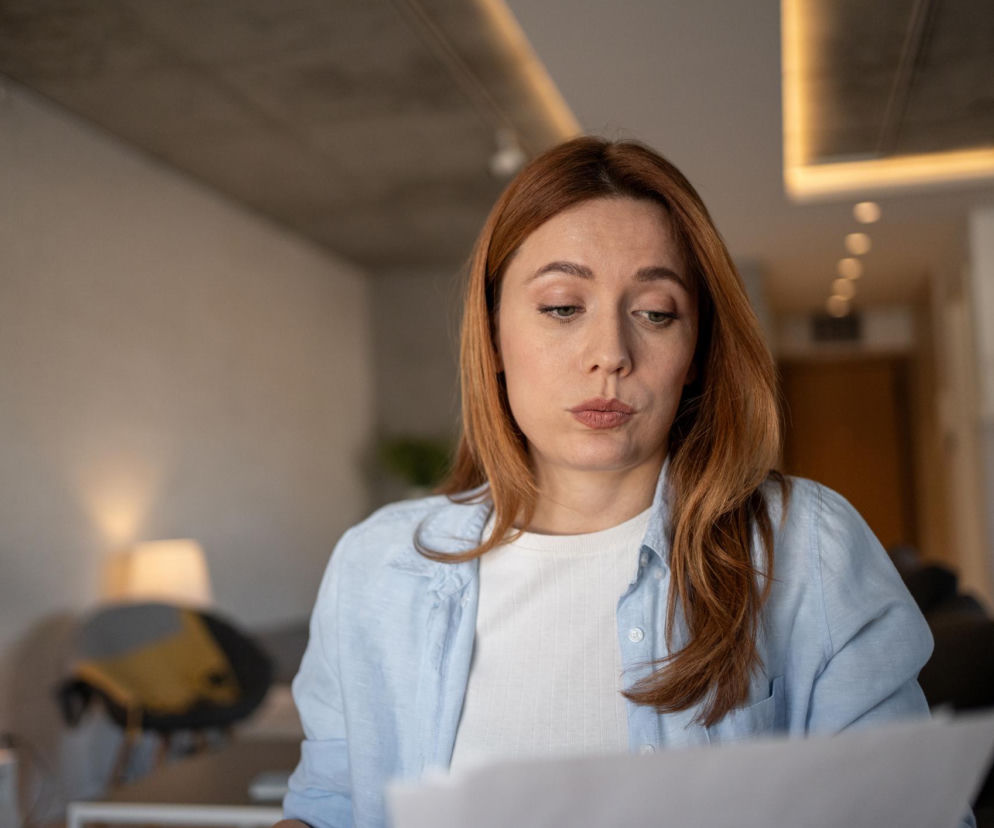Kobieta o rudych włosach, ubrana w białą bluzkę i błękitną koszulę, wpatruje się w kartki papieru, odzwierciedlając zjawisko jamais vu. W pomieszczeniu z lampą i meblami, symbolizuje moment, gdy znane słowo nagle wydaje się obce. Dowiedz się więcej o tym fenomenie na Poradnik Zdrowie.