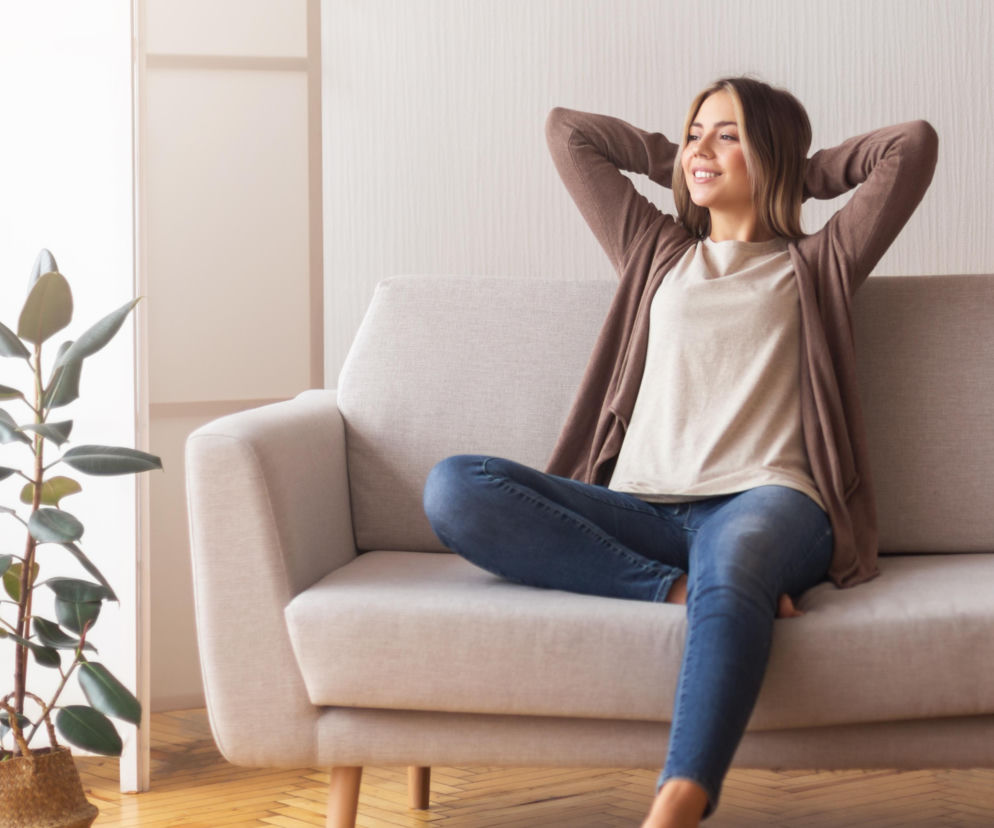 Kobieta w brązowym kardiganie i jeansach, siedząca z podwiniętą nogą na beżowej sofie, co jest szkodliwym nawykiem dla kręgosłupa i stawów, o czym przeczytasz więcej na Poradnik Zdrowie.