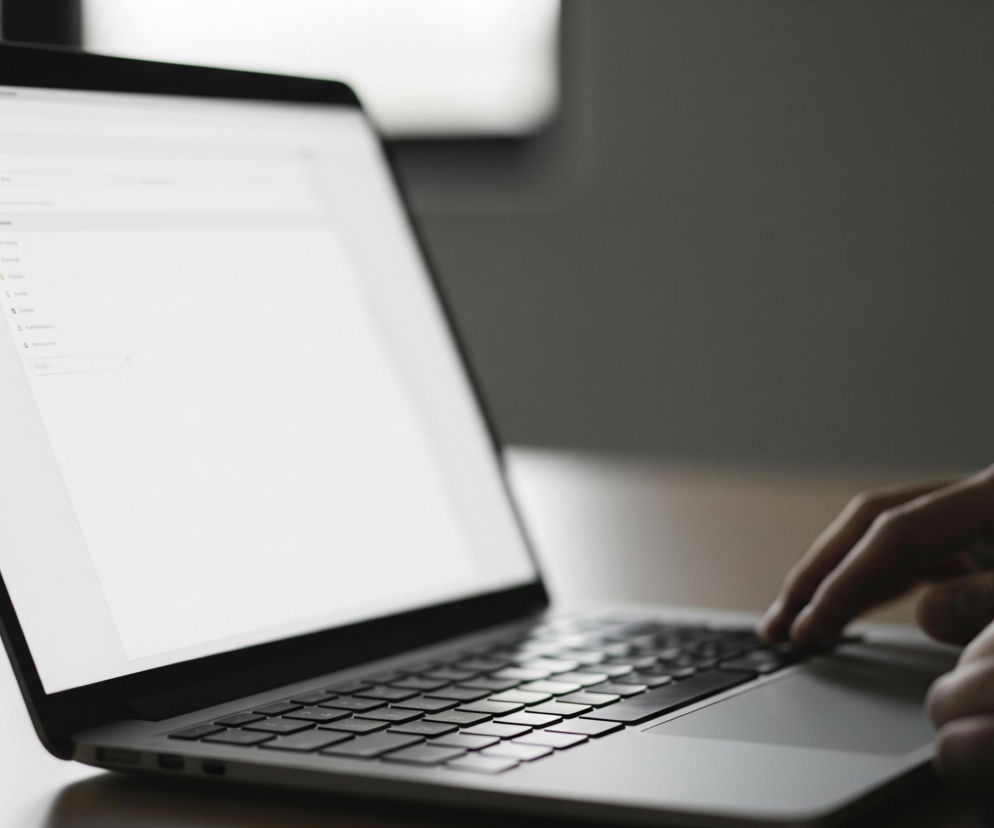 Na pierwszym planie, po lewej stronie kadru, znajduje się otwarty laptop w kolorze srebrnym z czarną klawiaturą i czarną ramką ekranu. Ekran laptopa jest jasny, biały, z częściowo widocznym interfejsem użytkownika przeglądarki internetowej lub aplikacji, zawierającym kilka ikon i tekst, ale w większości pusty. Po prawej stronie, w prawym dolnym rogu, widoczne są fragmenty dwóch rąk, z palcami spoczywającymi na klawiaturze laptopa, sugerujące pisanie lub nawigację. Tło jest rozmyte, ciemne, z niewielkim jasnym obszarem w górnej części kadru, sugerującym okno lub źródło światła.