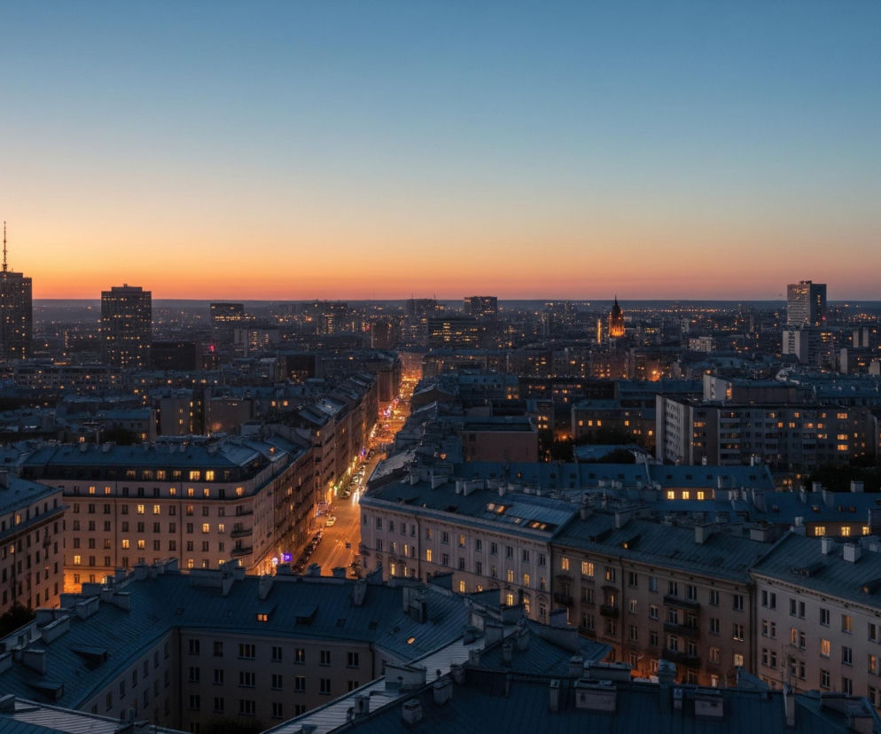 W oddali rozciąga się miejski krajobraz z licznymi budynkami, gdzie dominują odcienie szarości i granatu pod niebem przechodzącym z intensywnego pomarańczu zachodzącego słońca w dolnej części, do jasnego błękitu w górnej. W centrum widoczny jest długi, oświetlony od dołu pas ulicy z samochodami i drobnymi światłami w oddali. Na pierwszym planie znajdują się dachy budynków pokryte ciemnoniebieską blachą, z licznymi kominami i świetlikami, a w ich oknach jarzą się ciepłe, żółte światła. W oddali, po lewej stronie, wyróżnia się wysoki budynek z iglicą na szczycie.